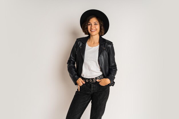 Photo de mode en studio d'une femme brune élégante en chapeau noir et veste en cuir posant sur blanc