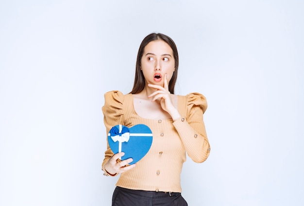 Photo d'un modèle de jeune femme tenant une boîte-cadeau en forme de coeur contre un mur blanc.