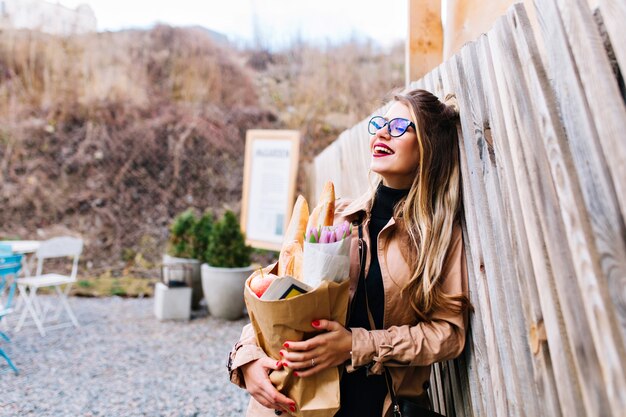 Photo à moitié tournée d'une jolie femme avec un sac en papier de délicieux repas du supermarché. Charmante fille transportant des aliments frais pour le déjeuner avec sa famille posant avec une expression de visage rêveur.