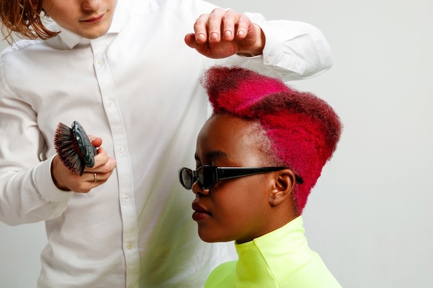 Photo montrant une femme afro-américaine au salon de coiffure. Photo de Studio de jeune fille gracieuse avec coupe courte élégante et cheveux colorés sur fond gris et mains de coiffeur.