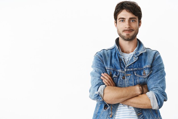 Photo non modifiée d'un bel homme adulte chaud et charismatique avec une barbe et des yeux bleus dans une veste en jean élégante sur un t-shirt rayé souriant optimiste, joyeux comme debout dans une pose confiante avec les mains croisées