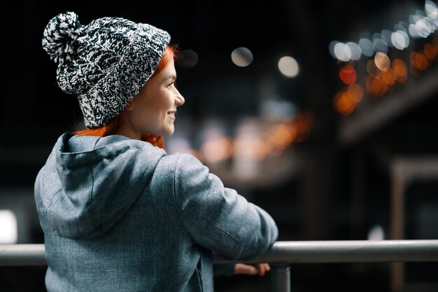 Photo de nuit en plein air d'une jeune belle fille souriante heureuse bénéficiant d'une décoration festive, dans la rue de la ville européenne, portant un bonnet tricoté