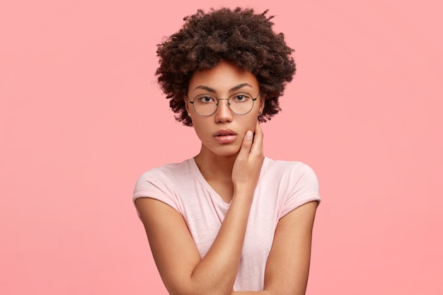 Photo d'une pigiste sérieuse en tenue décontractée, regarde mystérieusement, garde la main sur le cou, contemple une nouvelle stratégie de travail, pose contre un mur rose. Les gens et le style de vie