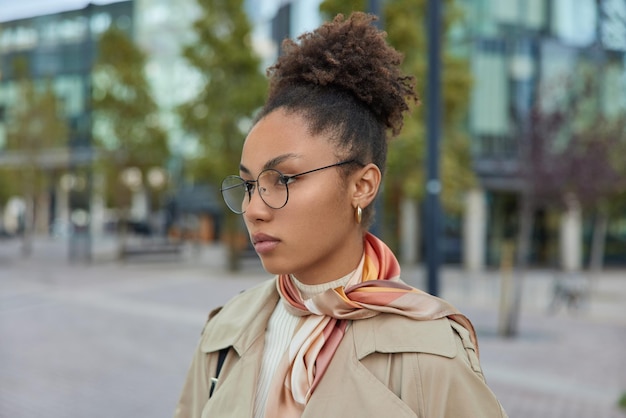 Photo en plein air d'une enseignante sérieuse et intelligente portant des lunettes rondes et des promenades en manteau dans la ville.