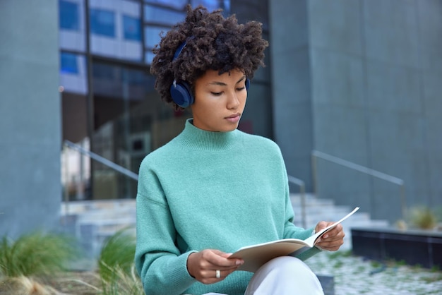 Photo en plein air d'une étudiante bouclée intelligente écoute une diffusion éducative dans des écouteurs et apprend des informations à partir d'un manuel à l'extérieur