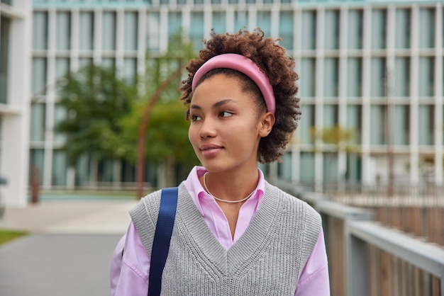 Photo gratuite photo en plein air d'une étudiante pensive aux cheveux bouclés qui revient de cours porte un bandeau et des vêtements décontractés soignés se promène à l'extérieur pose sur fond urbain flou concept de personnes et de style de vie