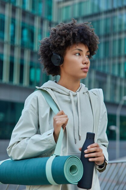 Photo en plein air d'une femme sportive aux cheveux bouclés tenant une bouteille d'eau portant un karemat roulé concentré porte des poses de sweat-shirt à l'extérieur