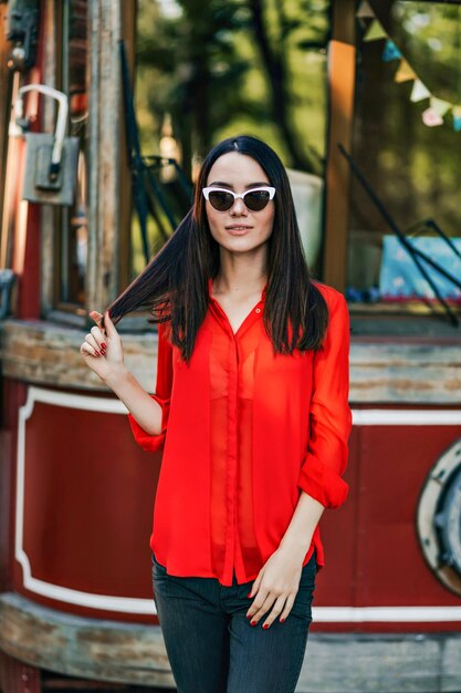 Photo en plein air d'une fille élégante et cool aux cheveux courts en chemise rouge tenant une tablette à l'extérieur Charmante femme élégante en lunettes de soleil posant sur le fond de la ville