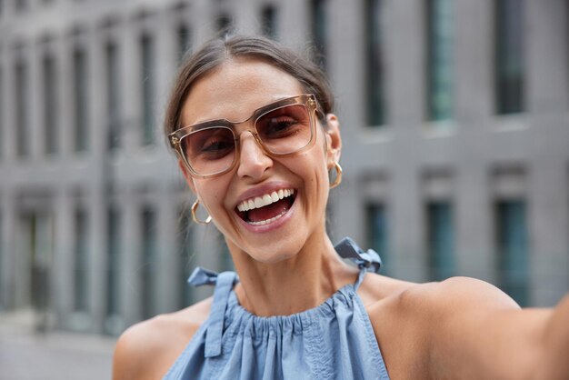 Une photo en plein air d'une jeune femme heureuse souriante portant des lunettes de soleil à la mode fait un selfie en se promenant dans des poses de ville à l'extérieur