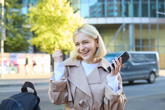 Photo gratuite photo en plein air d'une jeune femme joyeuse avec un smartphone à la main assise sur un banc de rue et se réjouissant