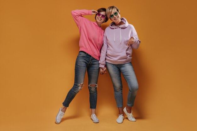 Photo pleine longueur de deux dames modernes portant des lunettes de soleil lumineuses, des sweats à capuche cool, des jeans skinny et des baskets souriant et se tenant la main sur fond orange.