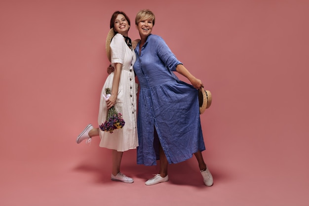 Photo pleine longueur de deux femmes aux cheveux courts et chapeaux de paille en robes midi et baskets blanches souriant et posant avec bouquet sur fond isolé.