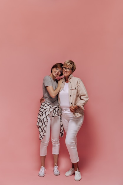 Photo pleine longueur de deux femmes avec une coiffure courte et cool et des lunettes modernes en pantalon blanc et des baskets souriant et étreignant sur fond isolé.