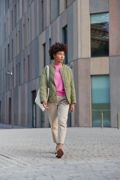 Photo gratuite une photo pleine longueur d'une femme aux cheveux bouclés porte un pantalon de veste et des baskets porte un sac en tissu se promène à l'extérieur dans le centre-ville près d'un bâtiment moderne atteint la destination nécessaire. mode de vie urbain