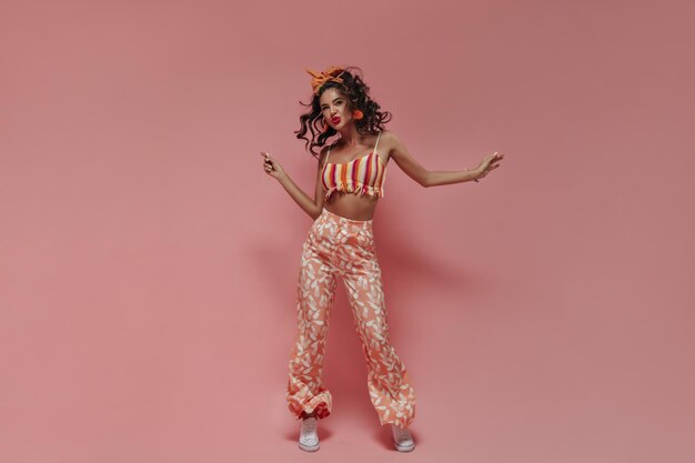 Photo pleine longueur d'une femme aux cheveux ondulés positive avec boucles d'oreilles et bandana dans un pantalon moderne rose à rayures et des baskets blanches dansant