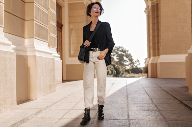 Photo gratuite photo pleine longueur d'une femme en pantalon léger, bottes et veste posant à l'extérieur. charmante femme avec sac à main noir et lunettes sourit à la rue.