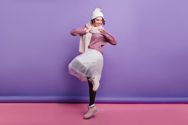 Photo pleine longueur d'une fille séduisante en jupe longue blanche posant sur une jambe et riant. portrait d'un magnifique modèle féminin en chaussures d'hiver et chapeau.