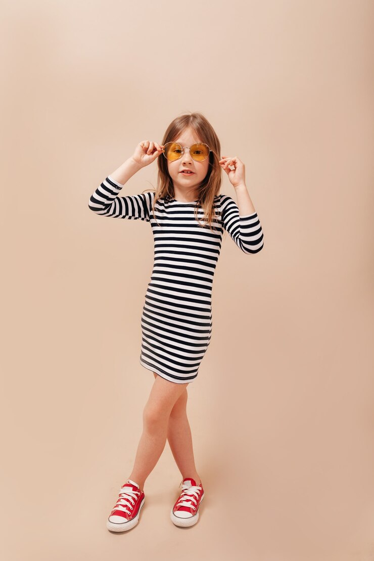 Photo pleine longueur de l'heureuse petite fille portant une robe dépouillée et des lunettes élégantes rondes s'amuse sur fond beige isolé