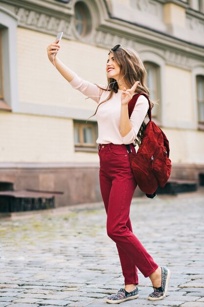 Photo pleine longueur de jolie fille aux cheveux longs faisant selfie-portrait au téléphone en ville. Elle a une couleur vineuse sur les vêtements et a l'air appréciée.