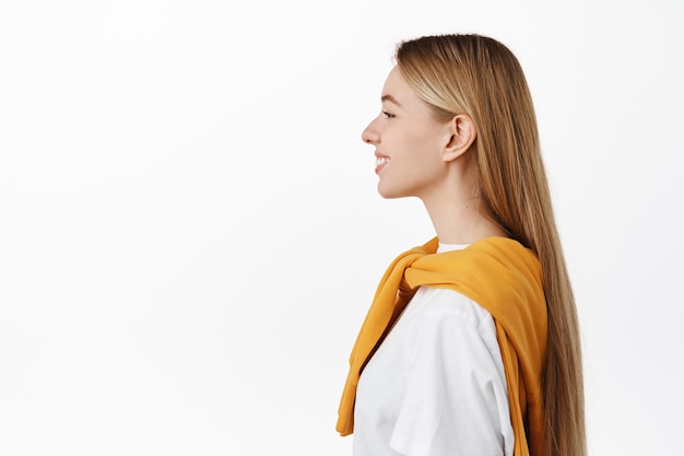 Photo gratuite photo de profil d'une belle femme heureuse avec de longs cheveux blonds raides, souriante joyeuse, regardant à gauche l'espace de copie, debout dans des vêtements décontractés contre un mur blanc