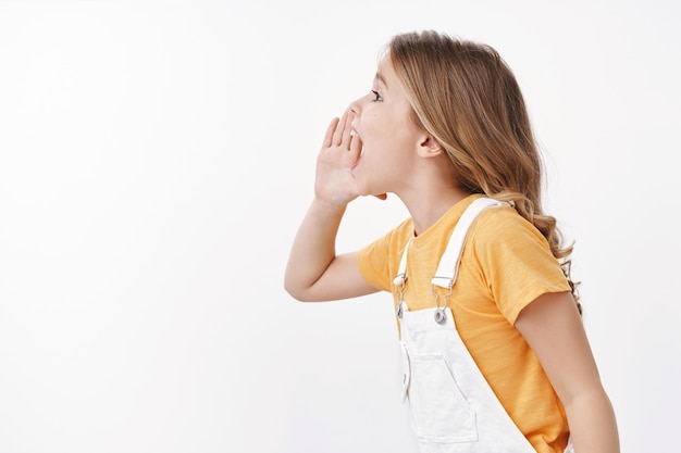 Photo gratuite photo de profil énergisée jolie petite fille charismatique enjouée, enfant appelant un ami, criant tenir la main près de la bouche ouverte, crier à haute voix le nom du parent, chercher le père, chercher quelqu'un, mur blanc