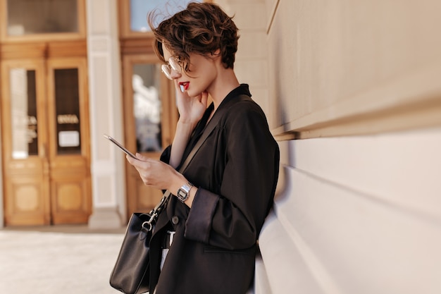 Photo de profil d'une femme cool aux cheveux courts en veste noire tient la tablette à l'extérieur. Belle femme dans des lunettes avec sac à main posant dans la rue.