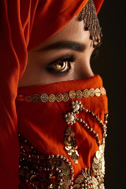 Photo de profil en gros plan d'une jolie jeune fille avec de beaux yeux charbonneux et de beaux bijoux sur le front, portant le hijab en terre cuite orné de paillettes. Elle pose de côté sur un fond sombre