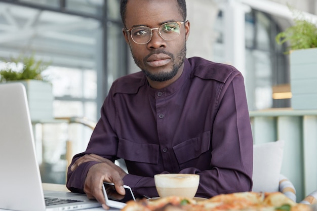 Photo recadrée d'un bel homme à la peau sombre porte des lunettes rondes et une chemise formelle, utilise les technologies modernes pour le travail