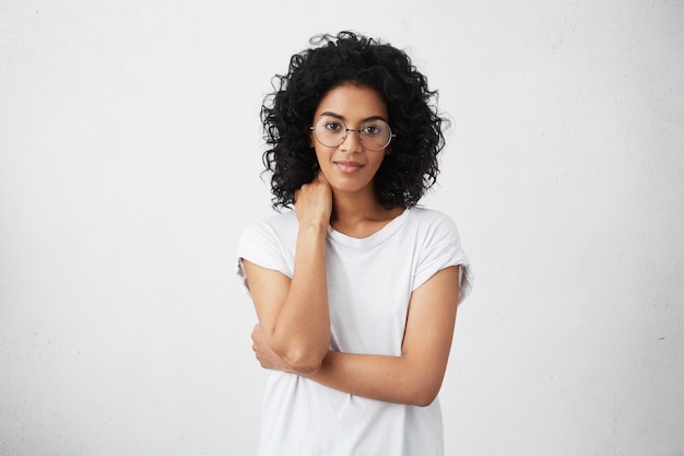 Photo recadrée de belle amicale jolie jeune femme afro-américaine souriante posant avec une posture fermée, légèrement souriante, ayant un regard timide, portant des lunettes Émotions et gestes humains