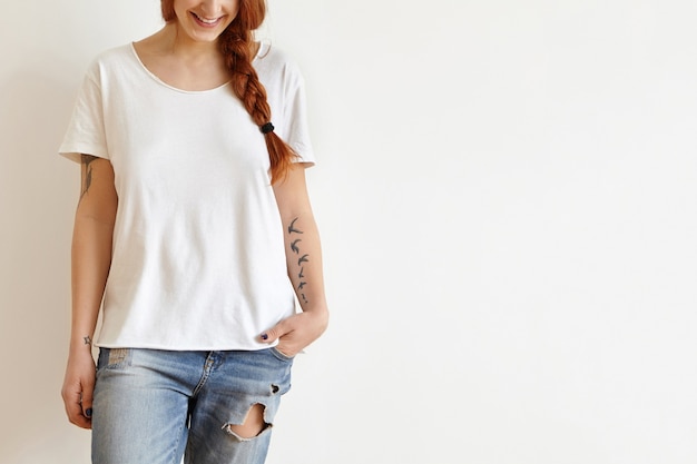 Photo recadrée de la belle jeune femme rousse élégante avec tresse et tatouages souriant joyeusement