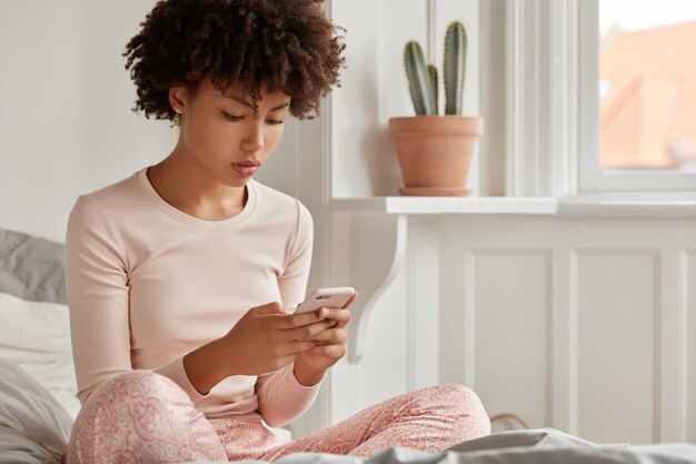 Photo recadrée d'une blogueuse jeune femme avec coupe de cheveux afro, habillée en pyjama, tient un téléphone intelligent