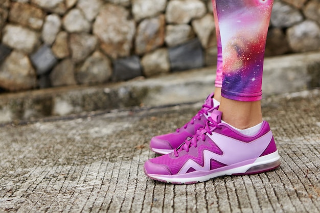 Photo gratuite photo recadrée d'une coureuse en forme portant des chaussures de course violettes et des leggings à imprimé spatial debout sur du béton de pierre tout en se préparant pour l'entraînement de jogging.