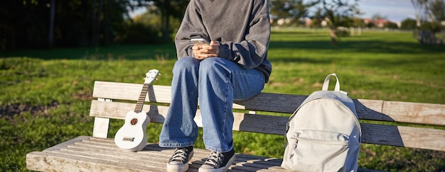 Photo gratuite photo recadrée du corps d'une adolescente assise sur un banc avec un ukulélé en utilisant un smartphone, les mains tenant un mobile