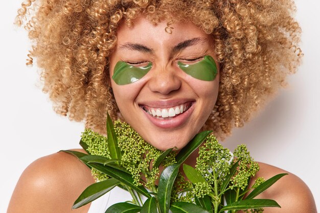 Une photo recadrée d'une femme aux cheveux bouclés ravie qui sourit largement garde les yeux fermés, des rires positifs tiennent positivement une plante verte fraîche applique des tampons d'hydrogel isolés sur fond blanc. Concept de peau d'oeil