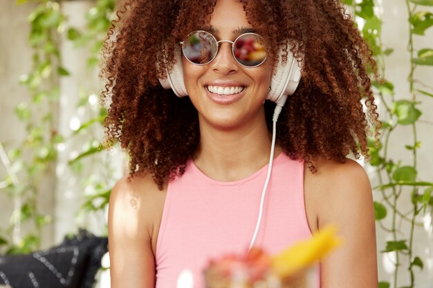 Photo recadrée d'une femme heureuse à la peau foncée avec une coiffure afro touffue écoute la playlist préférée dans les écouteurs, étant de bonne humeur, porte des lunettes de soleil à la mode et un t-shirt rose, a un sourire éclatant