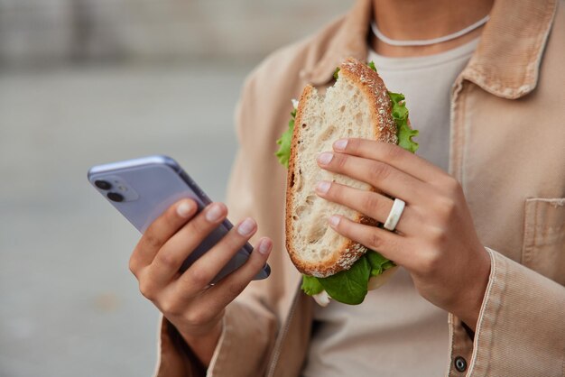 Photo recadrée d'une femme méconnaissable qui mange un délicieux sandwich et utilise un téléphone portable pour envoyer des SMS, vêtue de vêtements élégants, pose à l'extérieur sur fond flou. Concept de restauration rapide