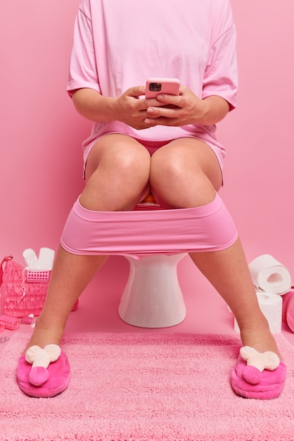 Photo gratuite photo recadrée d'une femme méconnaissable utilisant un smartphone alors qu'elle est assise sur la cuvette des toilettes porte une culotte de pantoufles tirée sur les jambes pose dans les toilettes accro aux technologies modernes mur rose