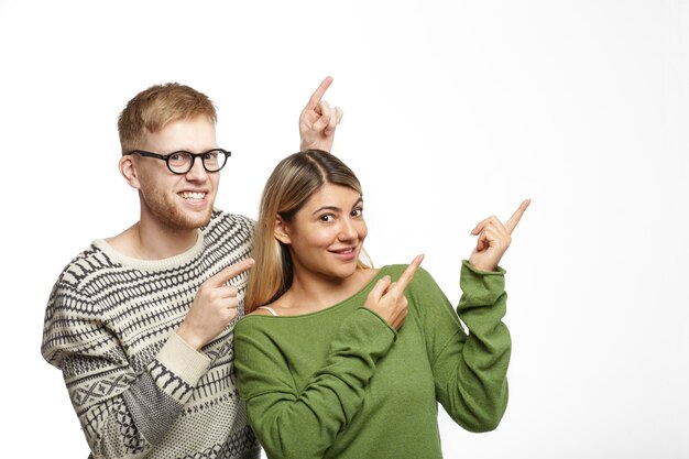 Photo recadrée d'heureux jeune couple marié homme et femme debout près l'un de l'autre, souriant joyeusement et pointant du doigt le mur de l'espace copie blanche pour votre publicité