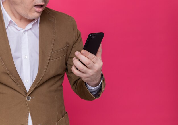 Photo recadrée d'un homme d'âge moyen en costume tenant un téléphone mobile en le regardant étonné