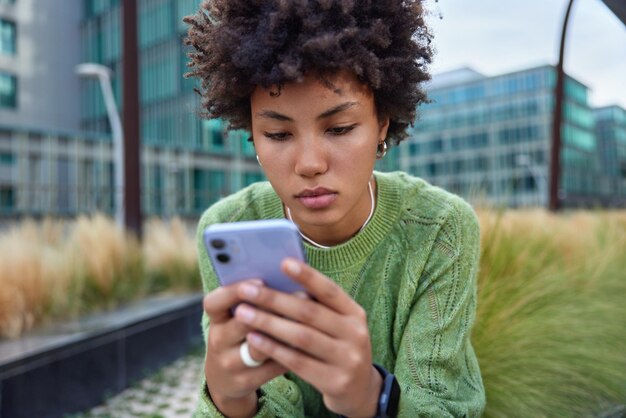 Photo recadrée d'une jeune femme sérieuse et séduisante concentrée sur des messages texte de type smatphone laisse des commentaires sous la publication dans des poses de blog à l'extérieur sur fond flou. Technologies modernes