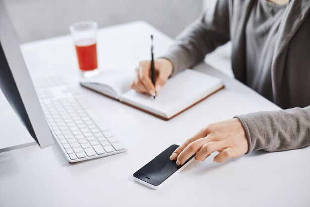 Photo recadrée de mains écrivant dans le bloc-notes et touchant le smartphone. Employé travaillant au bureau, vérifiant le courrier via un ordinateur et buvant du jus frais pour augmenter l'énergie. Les délais sont proches