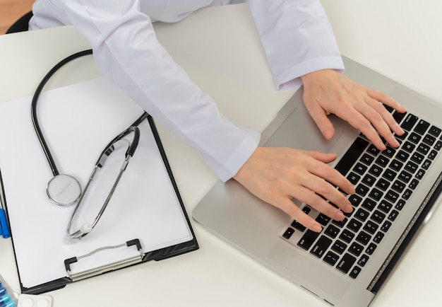 Photo recadrée de mains de femme médecin travaillant avec un ordinateur portable au bureau avec des outils médicaux et presse-papiers