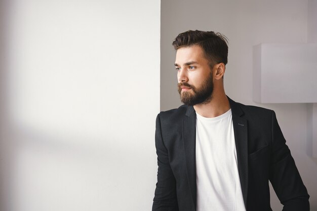 Photo recadrée de succès beau jeune homme mal rasé confiant portant des vêtements formels élégants posant près de la fenêtre, debout contre un fond de mur blanc blanc avec fond pour votre texte