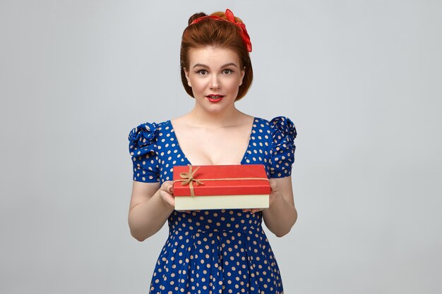 Photo de séduisante jeune femme de race blanche glamour portant maquillage lumineux et élégante robe à pois bleus, tenant une boîte rouge fantaisie, regardant la caméra, vous la remettant comme cadeau d'anniversaire