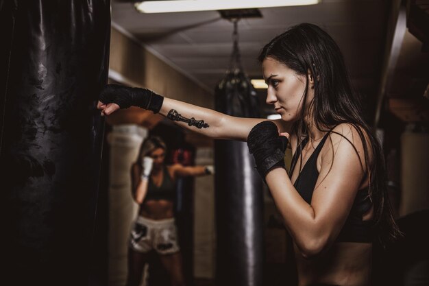 Photo sombre d'une jolie jeune femme dans une salle de sport sombre, qui s'entraîne à l'aide d'un sac de boxe.