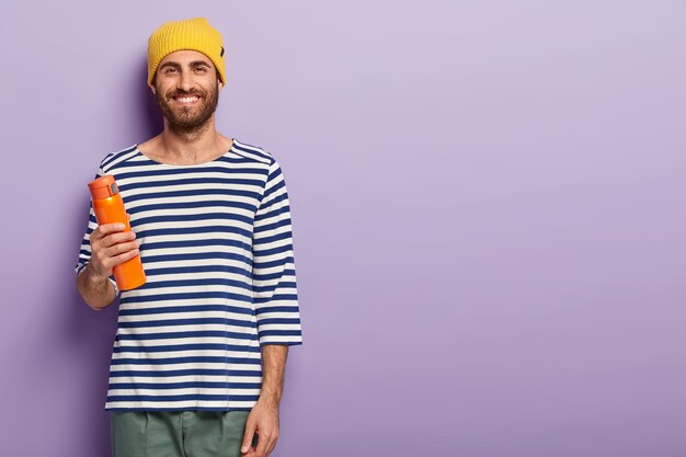 Photo de Studio de beau mec hipster souriant avec des poils foncés, porte un chapeau jaune et un pull rayé décontracté, tient une bouteille de boisson chaude, profite de temps libre, isolé sur un mur violet, espace libre à droite