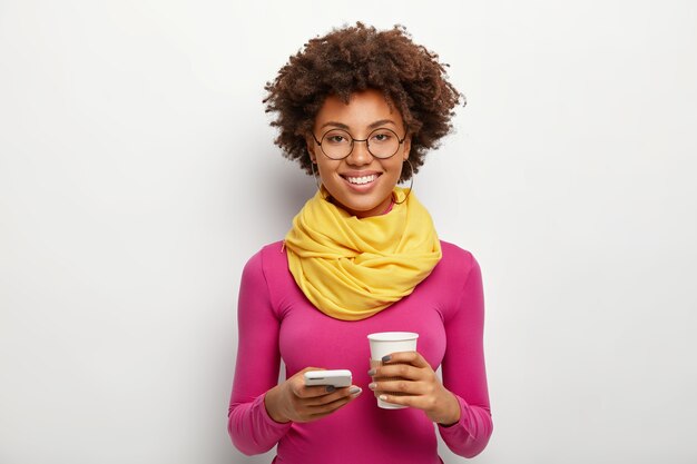 Photo de Studio de beau modèle féminin tient un téléphone portable moderne, fait de la planification dans un planificateur mobile, porte des lunettes, un col roulé rose et une écharpe, pose sur un mur blanc