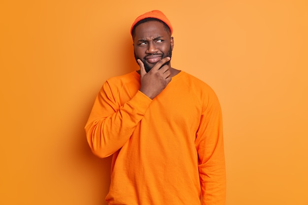 Photo gratuite photo de studio de bel homme barbu tient le menton regarde pensivement de côté pense profondément à quelque chose porte chapeau et pull pose contre un mur orange vif