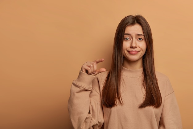 Photo de Studio d'une belle femme non impressionnée fait un geste de petite taille, démontre un objet minuscule, porte un sweat-shirt ample