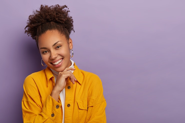 Photo gratuit photo de studio de charmante jeune femme heureuse avec une queue de cheval bouclée, touche doucement le menton, sourit joyeusement, porte une veste jaune à la mode, de grandes boucles d'oreilles rondes